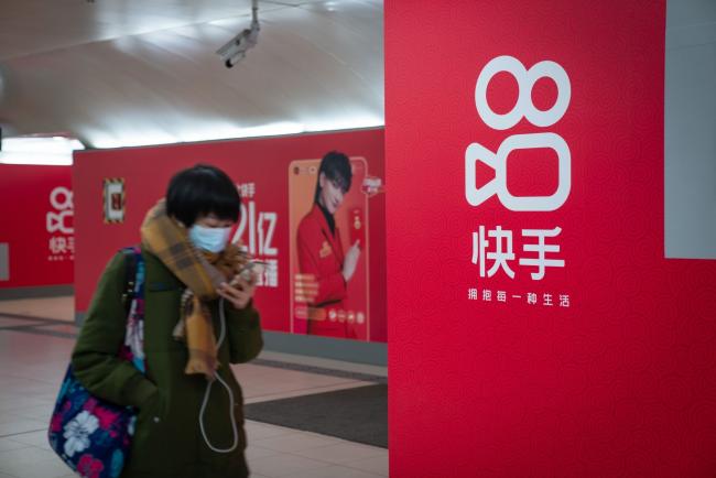 © Bloomberg. A passenger wearing a protective mask walks past Kuaishou Technology advertisements at a subway station in Beijing, China, on Wednesday, Feb. 3, 2021. Kuaishou Technology, the operator of China's most popular video service after ByteDance Ltd.'s Douyin, raised HK$42 billion ($5.4 billion) after pricing its Hong Kong initial public offering at the top of a marketed range. Photographer: Yan Cong/Bloomberg