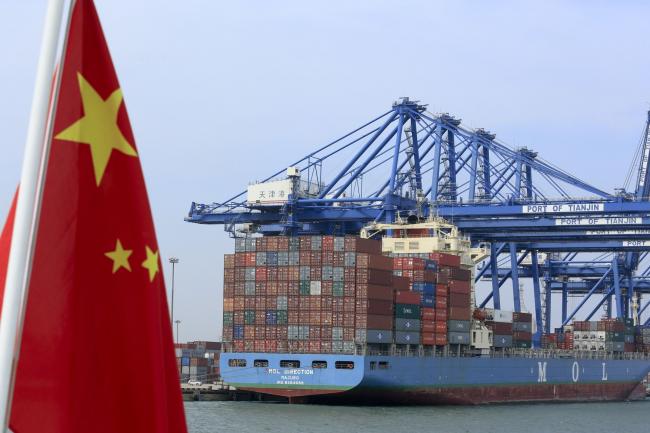 © Bloomberg. A Chinese flag flies on a vessel moving past shipping containers being unloaded at a Tianjin Port Group Co. dock in Tianjin, China. Photographer:Nelson Ching/Bloomberg 
