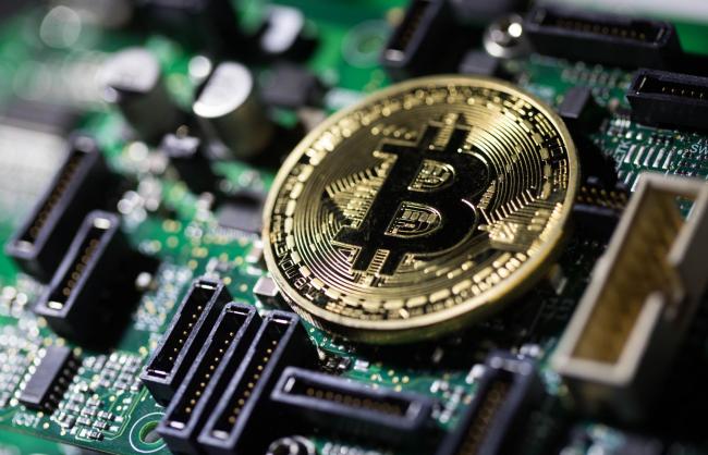 © Bloomberg. A coin representing Bitcoin cryptocurrency sits on a computer circuit board in this arranged photograph in London, U.K., on Tuesday, Feb. 6, 2018.  Photographer: Chris Ratcliffe/Bloomberg