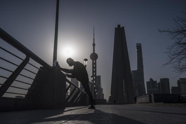 © Bloomberg. A man stretches on the Bund as skyscrapers of the Pudong Lujiazui Financial District stand across the Huangpu River during sunrise in Shanghai, China, on Friday, March 20, 2020. Most of China is now considered low risk and should return to normal work and life, Premier Li Keqiang said at a government meeting on the coronavirus, which is spreading rapidly in Europe, the U.S. and elsewhere. Photographer: Qilai Shen/Bloomberg