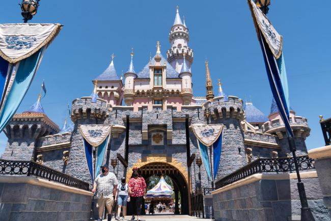 © Bloomberg. Guests wearing protective masks cross the bridge in front of Sleeping Beauty Castle during the reopening of the Disneyland theme park in Anaheim, California.