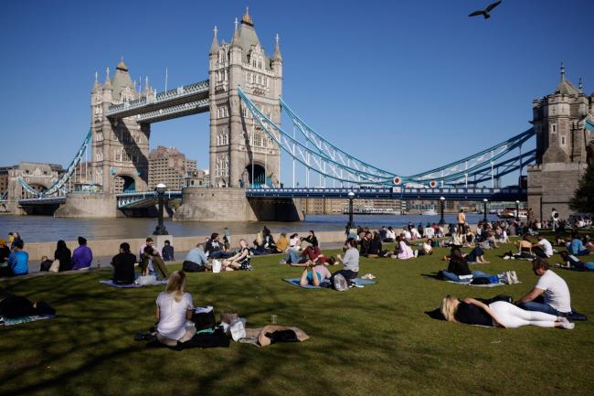 © Bloomberg. Pedestrians relax in the sunshine in a park on the south bank of the River Thames in view of Tower Bridge in London. Photographer: Jason Alden/Bloomberg
